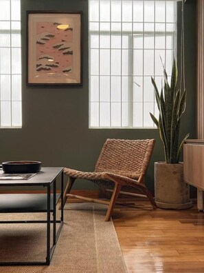 Une pièce comprenant une chaise en osier, une table avec un bol, une plante en pot et un tableau encadré accroché au mur.
