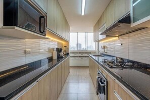 A modern kitchen with wooden cabinets, a black countertop, and a city view through the window. - Contemporary Pardo Miraflores Apartment By Geko (Miraflores)