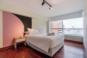A modern hotel room with a large bed, a bedside table, a window with a city view, and a curved wall design. - Contemporary Pardo Miraflores Apartment By Geko (Miraflores)