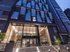 A modern hotel entrance with a glass facade, a staircase leading up to the entrance, and a visible outdoor area with plants. - Modern Dalias Miraflores Apartment By Geko (Miraflores)
