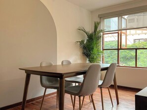 Un comedor con una mesa de madera y cuatro sillas, una planta grande en maceta y un ventanal con vista a follaje.