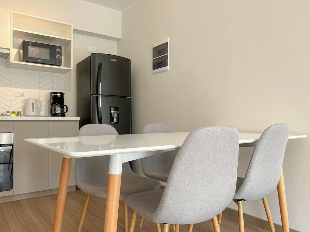 A modern kitchen with a white table, grey chairs, a black refrigerator, and a microwave on a white shelf.