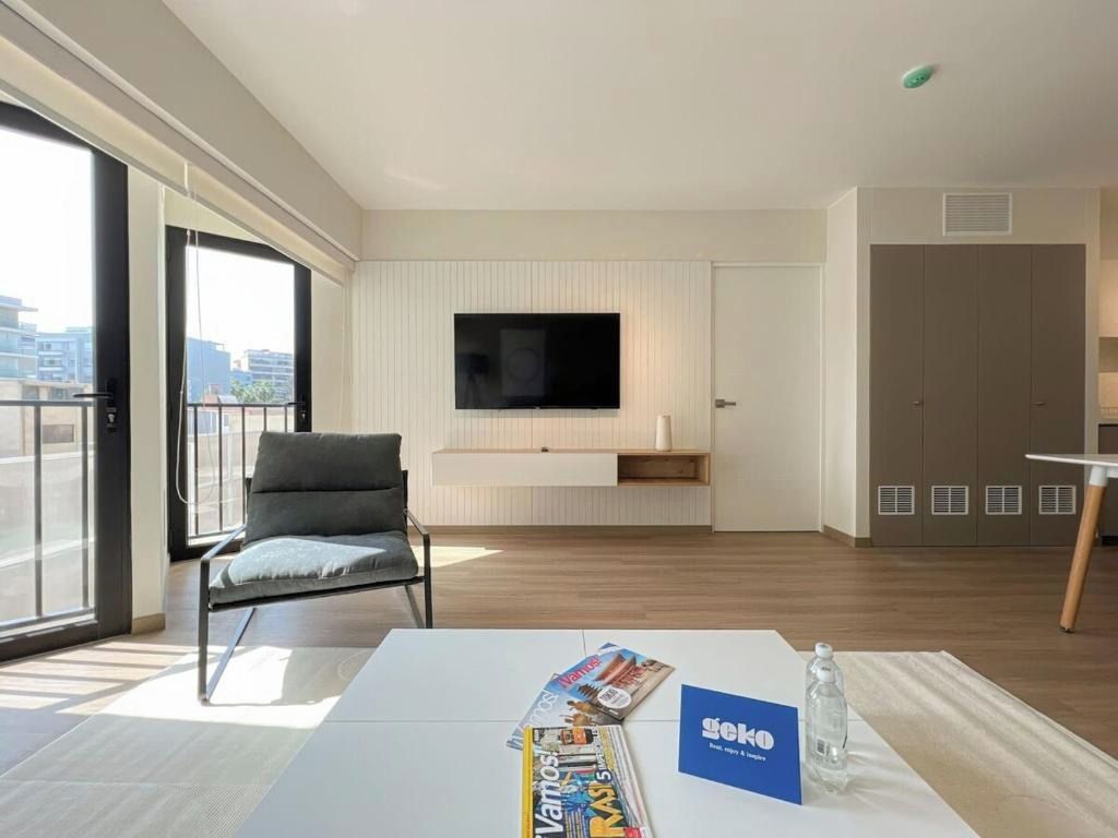 Ein modernes Hotelzimmer mit großem Fenster, einem an der Wand befestigten Flachbildfernseher, einem grauen Sofa, einem weißen Tisch mit Zeitschriften und einer Minibar.