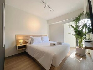 Ein modernes Hotelzimmer mit einem großen Bett, einem Nachttisch mit Lampe, einer Zimmerpflanze und Blick ins Freie.