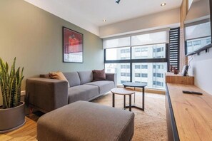 Una sala de estar moderna con un sofá, un reposapiés y una mesita. Hay una ventana grande con vistas a edificios.