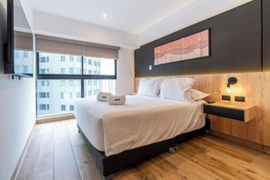 Ein modernes Hotelzimmer mit einem großen Bett, einem Holz-Nachttisch, einem an der Wand montierten Fernseher und einem Fenster mit Blick auf die Stadt.