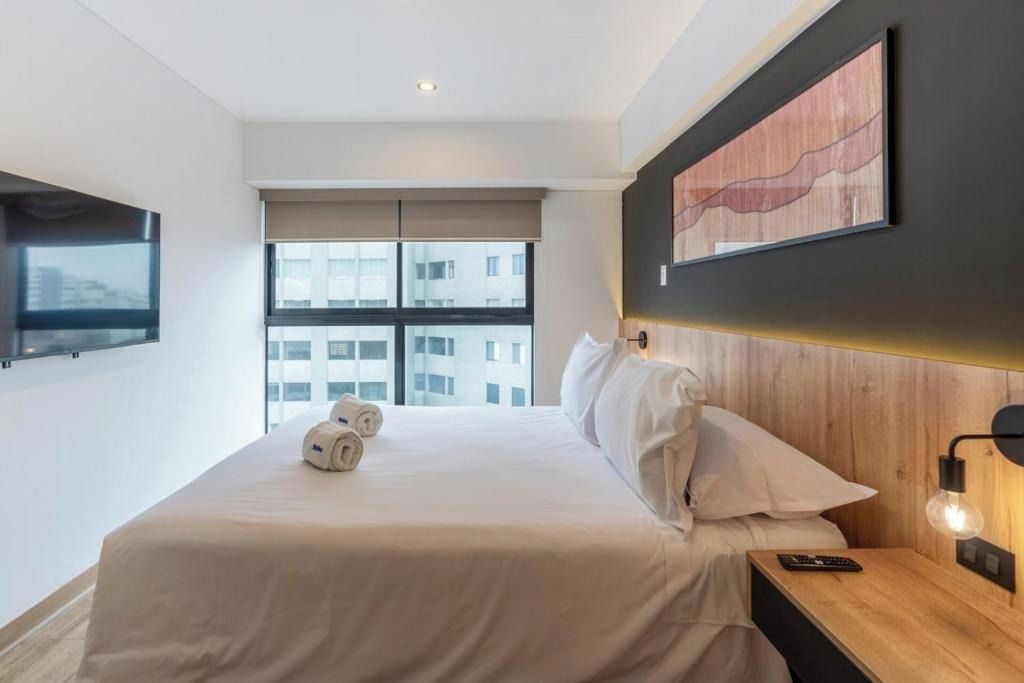 A hotel room with a large bed, a flat-screen TV mounted on the wall, a wooden headboard, and a window with a view of buildings.