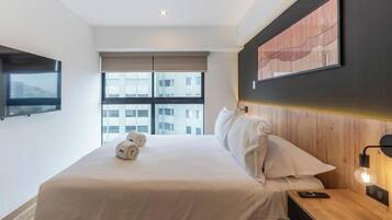 A hotel room with a large bed, a flat-screen TV mounted on the wall, a wooden headboard, and a window with a view of buildings.