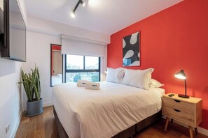 A modern bedroom with a bed, bedside table, lamp, potted plant, and a city view through the window. - Suárez Signature Apartment By Geko (San Isidro)
