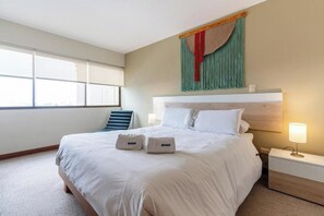 Ein Hotelzimmer mit einem großen Bett, einem Nachttisch mit Lampe, einem Fenster mit Jalousien und einem Sessel.