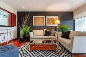 Ein modernes Wohnzimmer mit zwei Sofas, einem Couchtisch und einer Stehlampe. An den Wänden hängen zwei gerahmte Kunstwerke und es gibt Zimmerpflanzen.