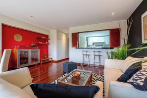 A modern living room with a red accent wall, a glass coffee table, and a kitchen area visible through an open door. - Coronel Portillo Residences By Geko (San Isidro)