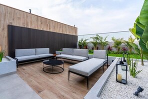 Ein moderner Außenbereich zum Sitzen mit einer Holzvertäfelung, grauen Sofas, einem runden Tisch und einem Pflanzgefäß mit Pflanzen.