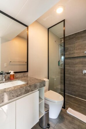 A modern bathroom with a glass shower enclosure, a white toilet, and a marble countertop with a sink. - Plaza 27 Residences By Geko (Miraflores)