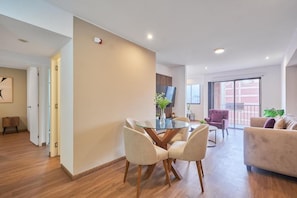A modern living room with a glass-top dining table, beige sofas, and a large window. - Modern 2Br Apartment With Pool, Gym & Coworking (Miraflores)