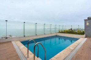 A rooftop swimming pool with a glass balustrade, a metal ladder, and a view of the city and sea. - Ejército City Lofts & Apartments By Geko (Miraflores)