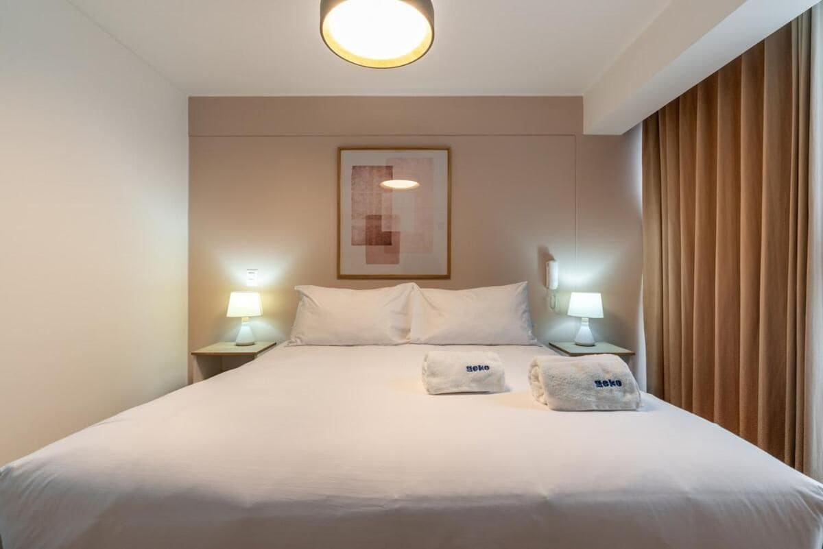 Quarto de hotel com uma cama de solteiro, duas luminárias de cabeceira, uma obra de arte emoldurada sobre a cama e uma janela com cortinas.