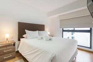 A hotel room with a large bed, a nightstand with a lamp, and a window with a view of buildings. - San Isidro Petit Thouars Living By Geko (San Isidro)
