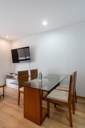 A modern dining area with a glass table, wooden chairs, a flat-screen TV mounted on the wall, and a small white cabinet. - San Isidro Petit Thouars Living By Geko (San Isidro)