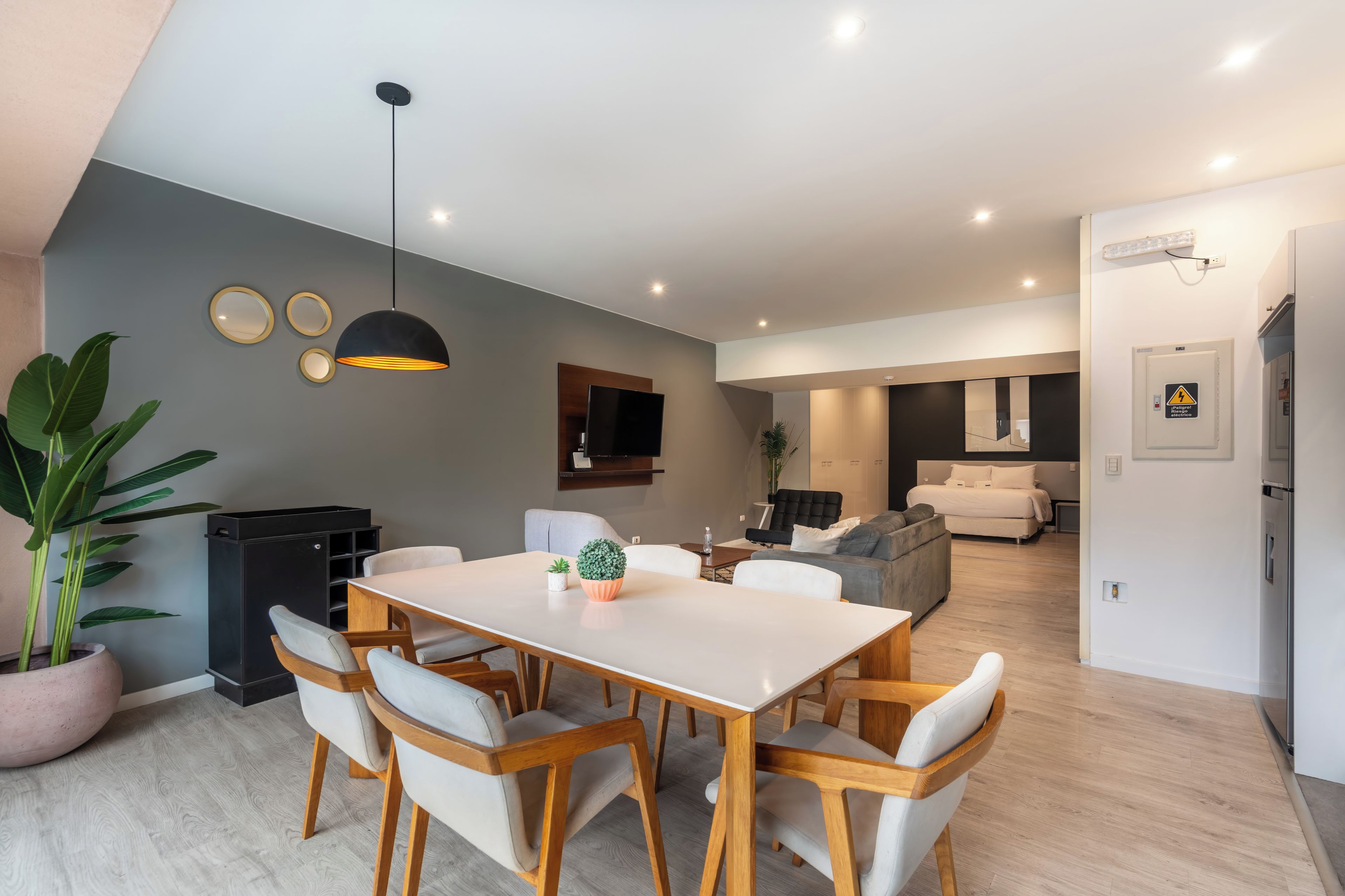 A modern living space with a dining area, a flat-screen TV mounted on the wall, a sofa, and a bed in the background.