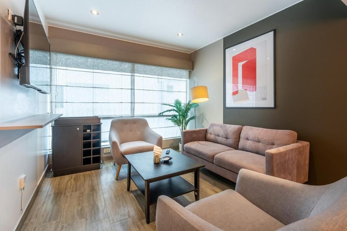 Una sala de estar moderna con un sofá, un sillón y una mesa de centro. Hay un televisor montado en la pared y una ventana con persianas.