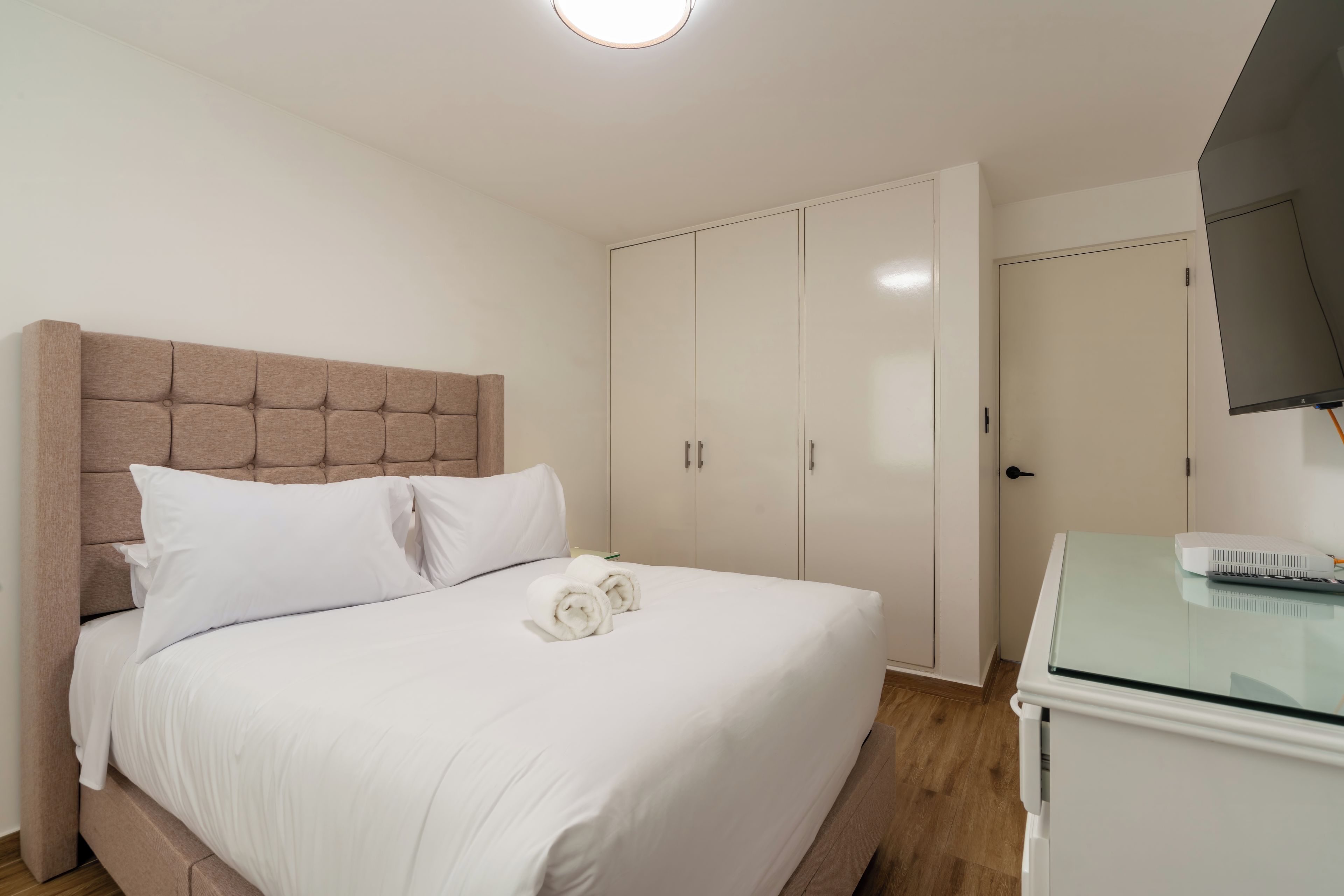 A neatly made bed with white linens, a headboard, and two rolled towels. There is a wardrobe with a mirror and a television on a stand.