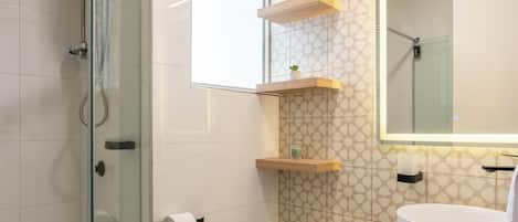A modern bathroom with a glass shower enclosure, a toilet, a sink with a marble countertop, and a mirror with a wooden shelf above it.