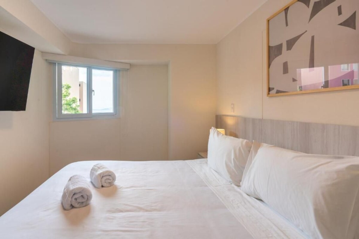 A hotel room with a bed, two rolled towels, a window, a wall-mounted TV, and a painting.