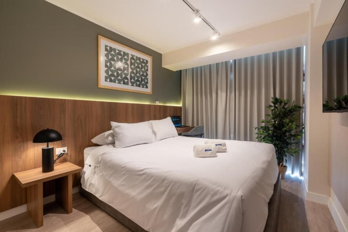 A modern hotel room with a bed, bedside table, lamp, and a large window with curtains.
