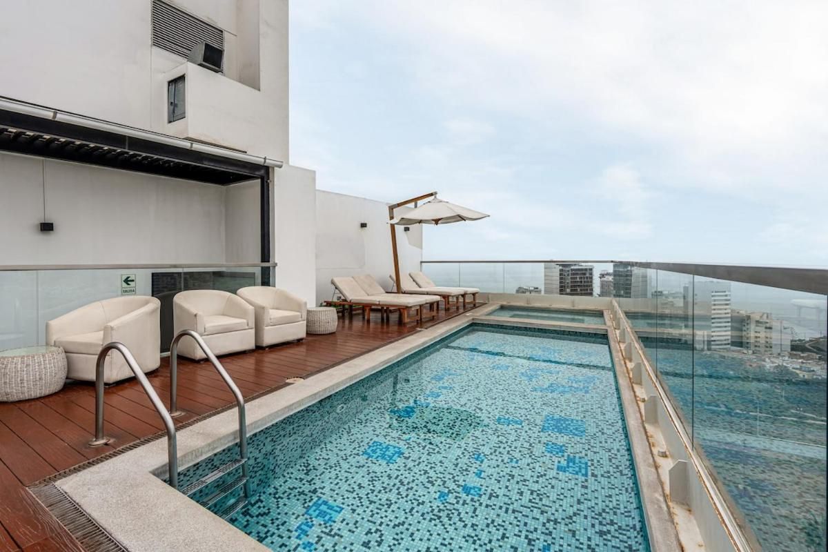 A rooftop pool area with lounge chairs and a glass balustrade overlooking a cityscape.