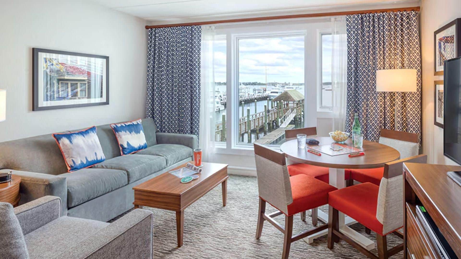 A modern hotel room with a grey sofa, a wooden coffee table, a dining table with red chairs, and a large window with a view of a marina.