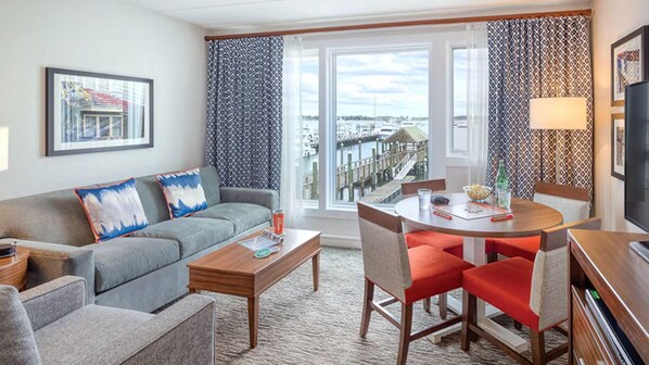 A modern hotel room with a grey sofa, a wooden coffee table, a dining table with red chairs, and a large window with a view of a marina.