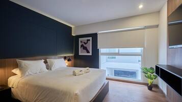 A modern hotel room with a large bed, a flat-screen TV, a potted plant, and a window with a view.