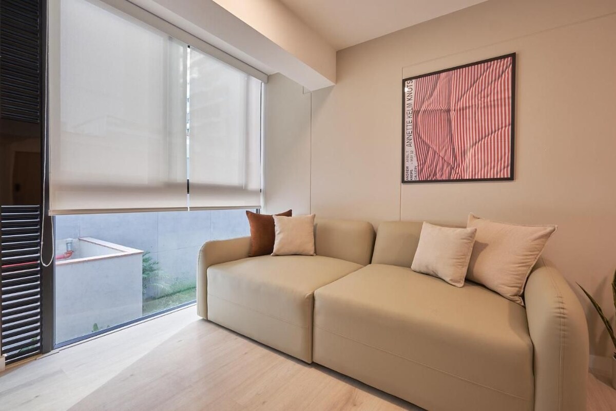 A modern living room with a beige sofa, a large window, and a framed artwork on the wall.