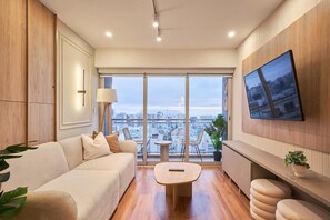 A modern living room with a sofa, a coffee table, a TV mounted on the wall, and a view of the city through large windows. - Pardo 510 Suites By Geko (Miraflores)