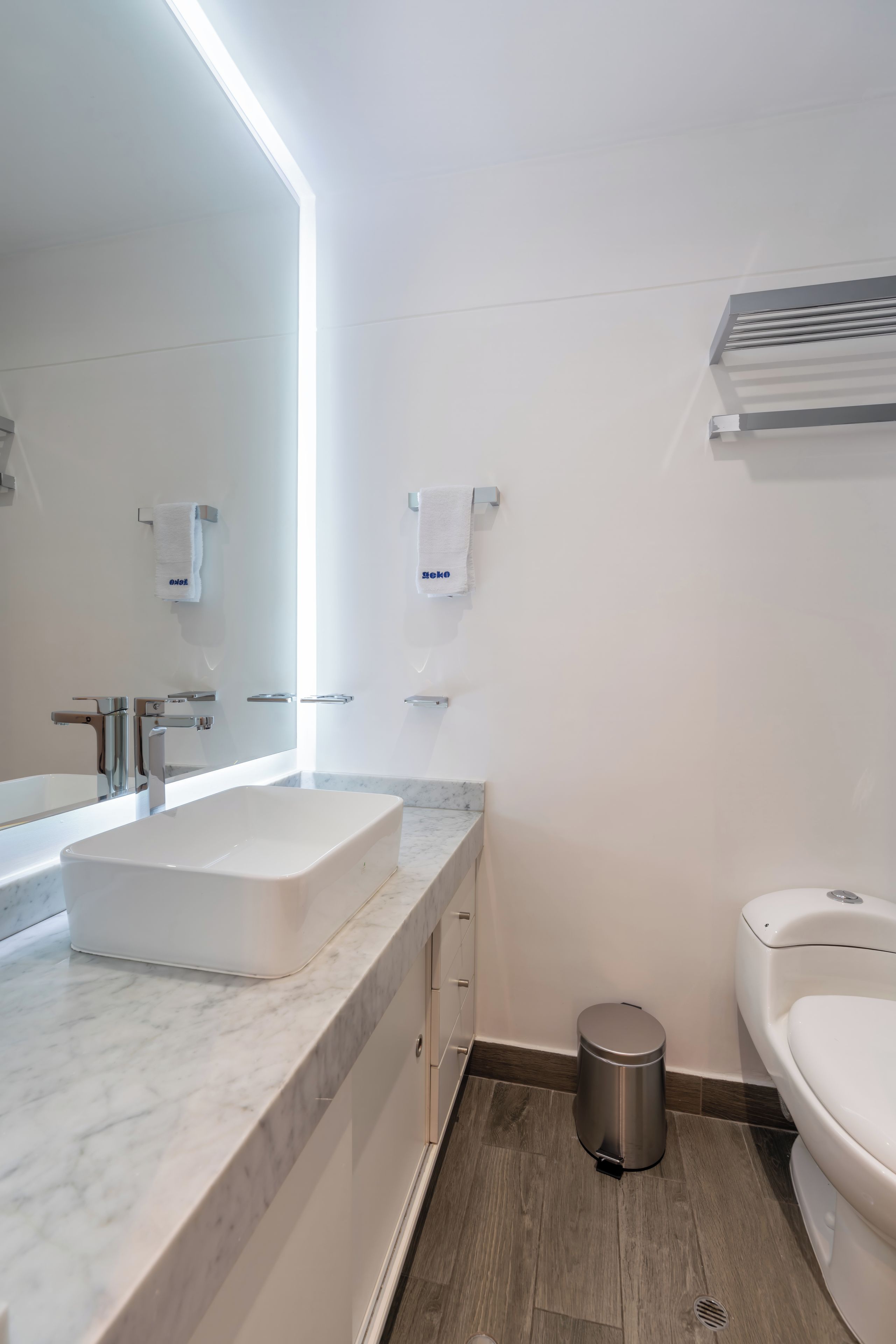 A modern bathroom with a marble countertop, a large mirror, a toilet, and a trash bin.