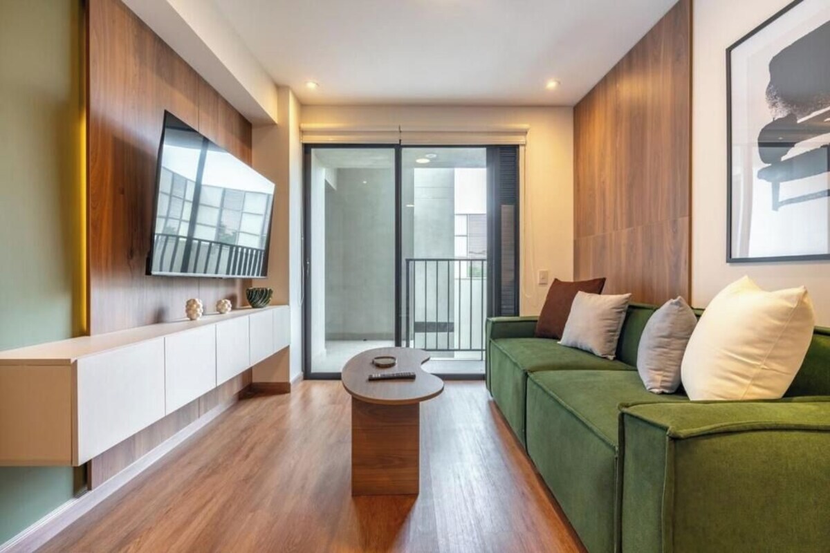 Ein modernes Wohnzimmer mit einem grünen Sofa, einem Holztisch, einem an der Wand montierten Fernseher und einer Glastür zum Balkon.