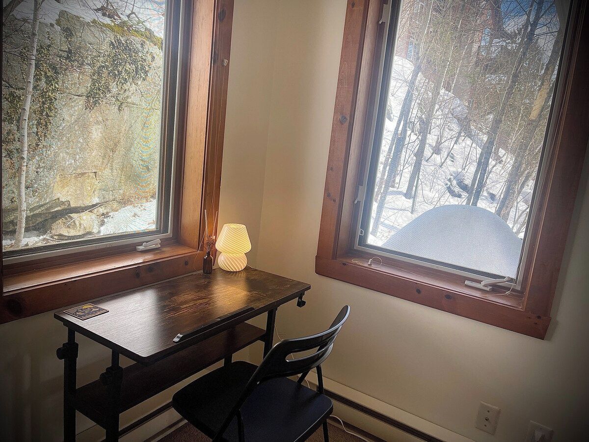 A room with a wooden table and chair, a lamp, and two windows with views of trees and a snowy landscape.