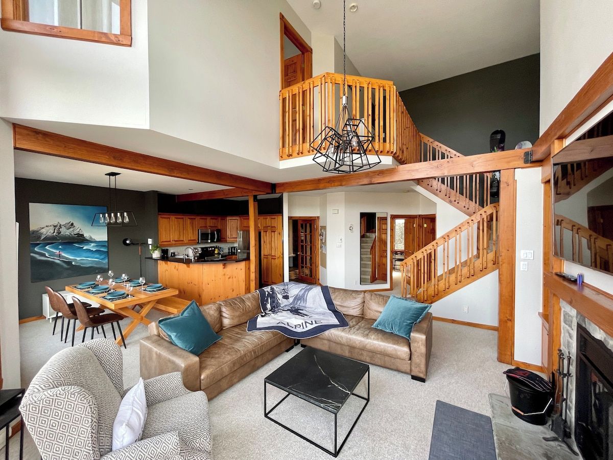 A spacious living area with a high ceiling, a wooden staircase, and a dining area with a view of the kitchen.