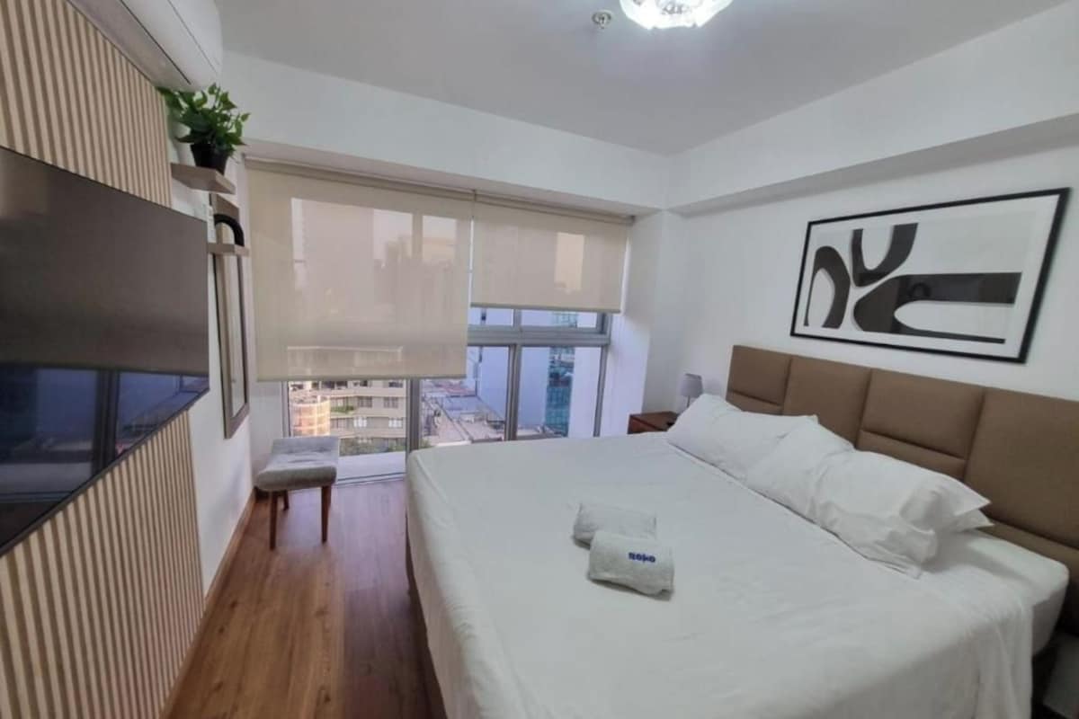 A hotel room with a large bed, a TV mounted on the wall, a window with a view of buildings, and a plant on a shelf.