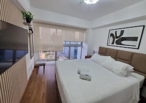 A hotel room with a large bed, a TV mounted on the wall, a window with a view of buildings, and a plant on a shelf. - Javier Prado 595 Residences By Geko (San Isidro)