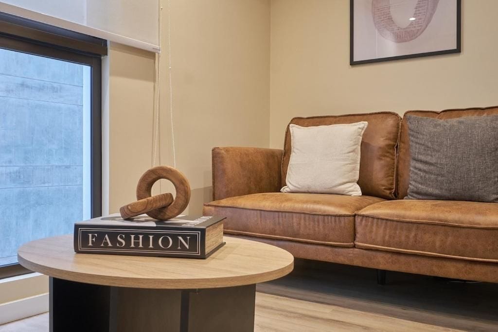 Ein braunes Ledersofa mit grau-weißem Kissen, ein runder Holztisch mit einem Buch mit dem Titel 'FASHION' und ein gerahmtes Bild an der Wand.