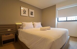A hotel room with a large bed, two bedside tables, and a window with blinds. - Work And Living Suites In Miraflores By Geko (Miraflores)