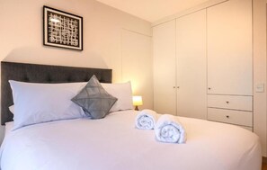 Habitación de hotel con cama, almohadas, una toalla doblada, mesita de noche con lámpara y armario.