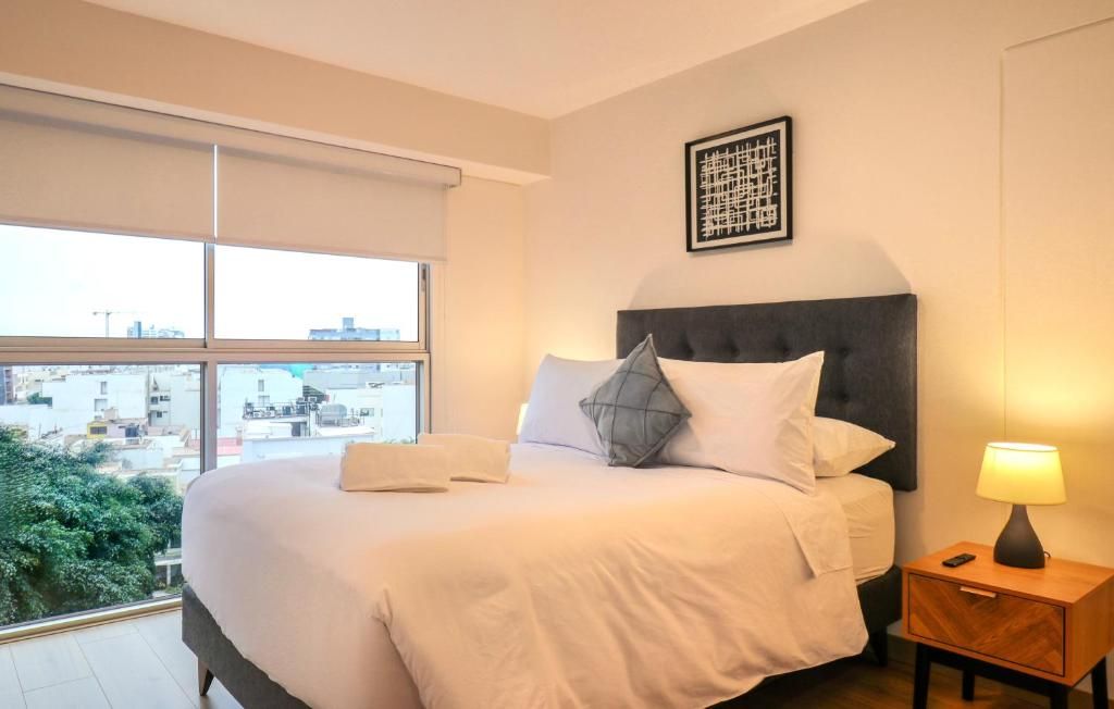 Habitación de hotel con una cama grande, una mesita de noche con una lámpara y una ventana con vistas a edificios y zonas verdes.