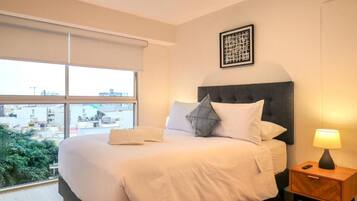 Habitación de hotel con una cama grande, una mesita de noche con una lámpara y una ventana con vistas a edificios y zonas verdes.