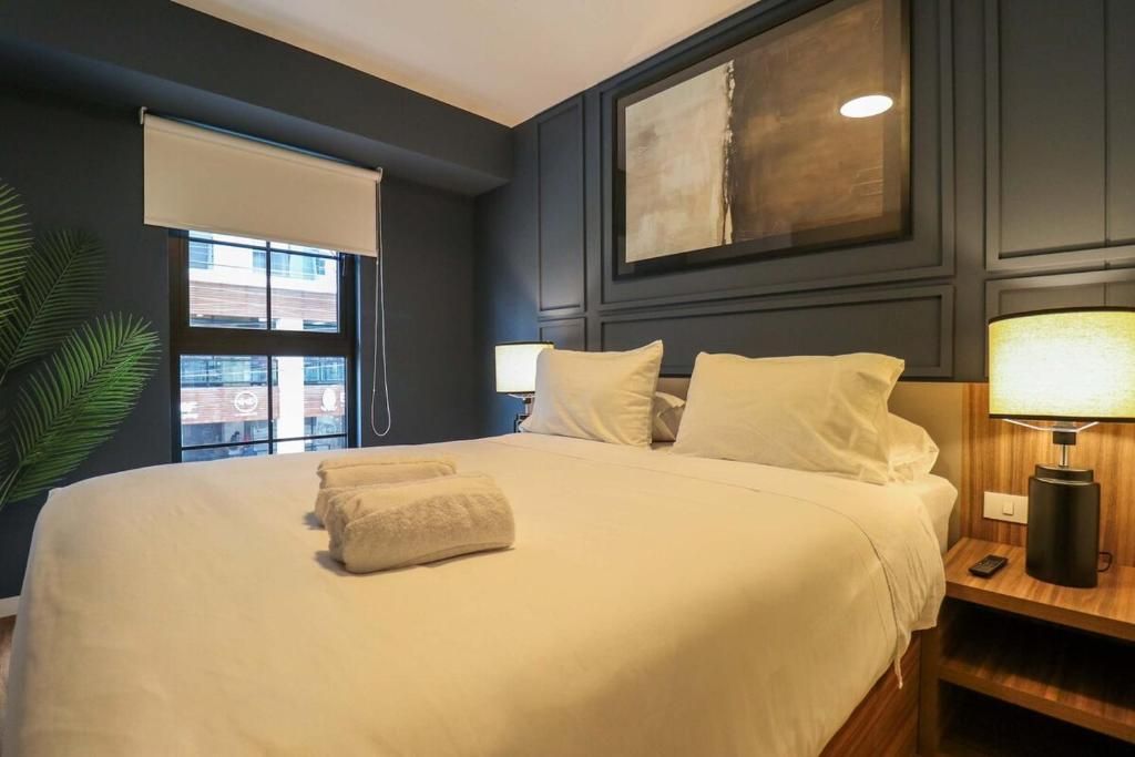 A hotel room with a large bed, a bedside table with a lamp, a window with a blind, and a framed artwork on the wall.