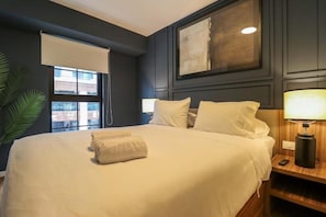 A hotel room with a large bed, a bedside table with a lamp, a window with a blind, and a framed artwork on the wall. - José Toribio Polo Residences By Geko (San Isidro)