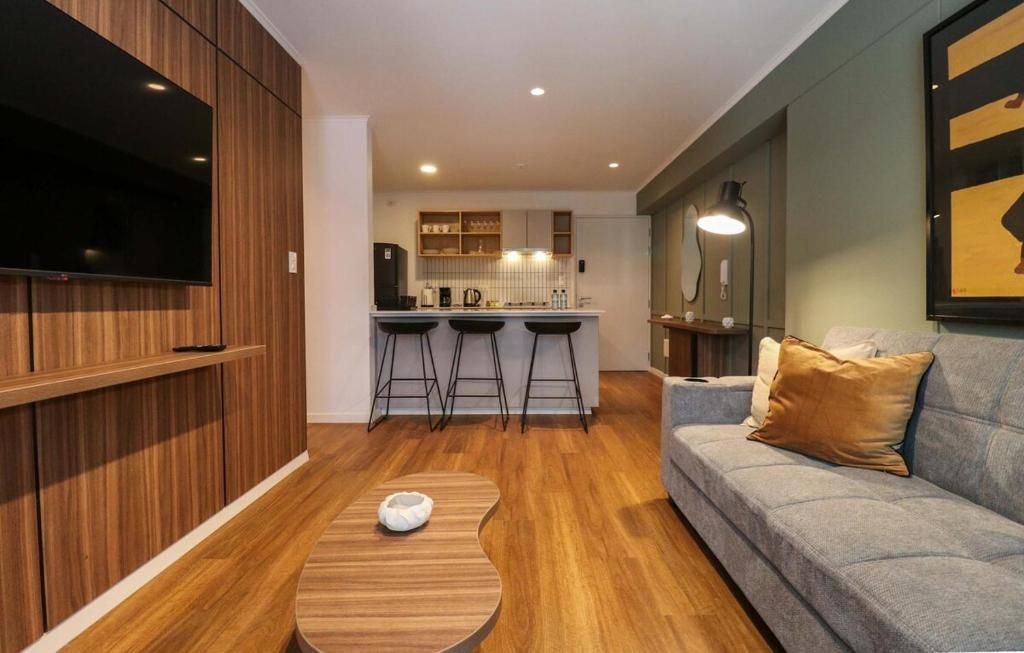 A modern living room with a grey sofa, wooden flooring, a TV mounted on a wooden panel, and a kitchen area with stools and a bar counter.