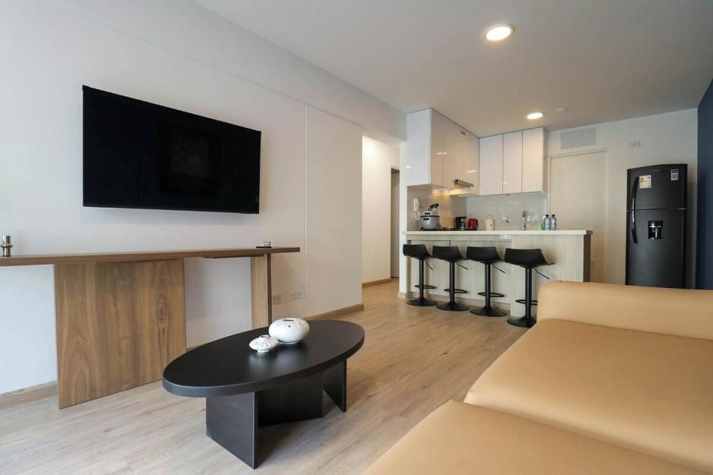 Una sala de estar moderna con un sofá, una mesa de centro, un televisor montado en la pared y una zona de cocina con un refrigerador y taburetes de bar.